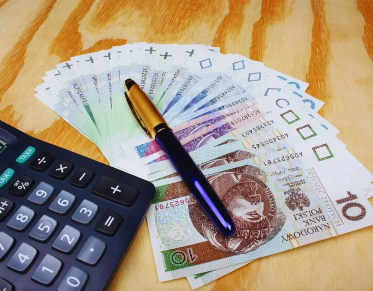 Obrazek wyróżniający do artykułu - wsparcie finansowe do zatrudnienia osoby niepełnosprawnej. Na zdjęciu widoczny jest wachlarz banknotów polskich złotych, rozłożony na drewnianym blacie. Banknoty o nominałach 10 złotych są starannie rozłożone w półokręgu. Na banknotach leży elegancki długopis o ciemnoniebieskim korpusie i złotej końcówce. Obok, po lewej stronie, znajduje się kalkulator z dużymi przyciskami, sugerujący, że obliczenia lub kalkulacje finansowe mogą być częścią tematu zdjęcia. Całość nawiązuje do kwestii finansów lub wsparcia pieniężnego.