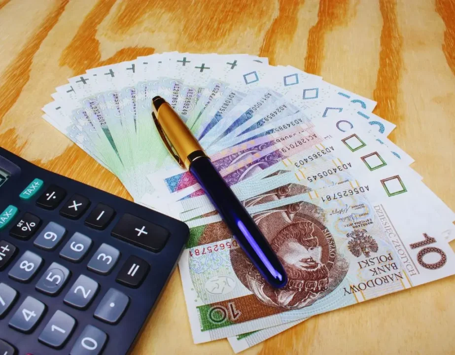 Na jakie wsparcie finansowe możesz liczyć, jeśli zatrudnisz osobę niepełnosprawną? 4 Obrazek wyróżniający do artykułu - wsparcie finansowe do zatrudnienia osoby niepełnosprawnej. Na zdjęciu widoczny jest wachlarz banknotów polskich złotych, rozłożony na drewnianym blacie. Banknoty o nominałach 10 złotych są starannie rozłożone w półokręgu. Na banknotach leży elegancki długopis o ciemnoniebieskim korpusie i złotej końcówce. Obok, po lewej stronie, znajduje się kalkulator z dużymi przyciskami, sugerujący, że obliczenia lub kalkulacje finansowe mogą być częścią tematu zdjęcia. Całość nawiązuje do kwestii finansów lub wsparcia pieniężnego.