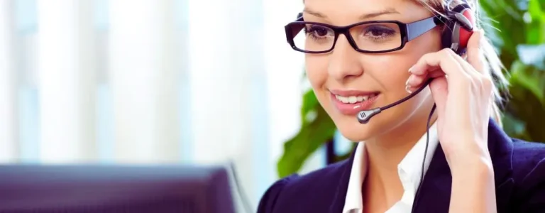 Obrazek do artykułu - zatrudnienie niewidomych w obsłudze klienta. Na zdjęciu widzimy kobietę w profesjonalnym stroju, która pracuje jako konsultantka telefoniczna. Kobieta ma na sobie ciemny garnitur oraz białą koszulę, co sugeruje formalne otoczenie pracy, takie jak biuro lub centrum obsługi klienta. Na głowie nosi słuchawki z mikrofonem, które pozwalają jej prowadzić rozmowy telefoniczne. Ma blond włosy związane do tyłu oraz okulary w ciemnych oprawkach. Kobieta uśmiecha się delikatnie, co sugeruje, że prowadzi przyjazną rozmowę z klientem. W tle widać rozmazane elementy biura i roślinność, co tworzy przyjazne i profesjonalne środowisko pracy. Zdjęcie pokazuje typową scenę pracy konsultantki obsługującej klientów.