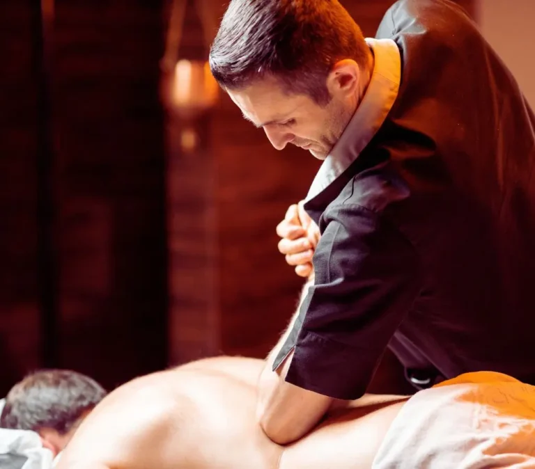 Obrazek do artykułu - zatrudnienie niewidomych w terapii. Na zdjęciu widzimy mężczyznę wykonującego masaż. Mężczyzna jest ubrany w ciemny strój, charakterystyczny dla terapeutów masażu. Skupia się na pracy, a jego postawa i gesty wskazują na intensywną technikę masażu - wykonuje masaż, używając przedramienia, aby wywrzeć większy nacisk na ciało osoby leżącej. Osoba masowana leży na brzuchu, z głową skierowaną w bok, a jej plecy są odsłonięte. Otoczenie jest ciepłe i przytulne, co tworzy odpowiednią atmosferę do relaksu. W tle widoczne są ciepłe brązowe odcienie, które sugerują, że miejsce to jest zaprojektowane do wyciszenia i odprężenia.