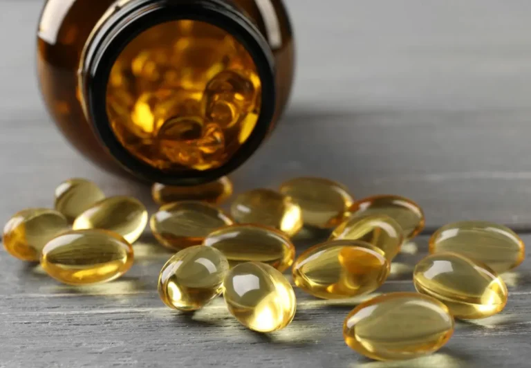 Na zdjęciu widzimy brązową butelkę z szerokim otworem, z której wysypały się kapsułki o złocistym, przezroczystym wyglądzie. Kapsułki mają owalny kształt, wyglądają na żelowe, co sugeruje, że mogą zawierać olej lub witaminy, często kojarzone z suplementami, takimi jak tran lub witamina D. Kapsułki rozłożone są na gładkiej, szarej powierzchni, a ich przejrzysta struktura pozwala dostrzec ciecz lub olej wewnątrz. Całość prezentuje się estetycznie i sugeruje tematykę zdrowia lub suplementacji.