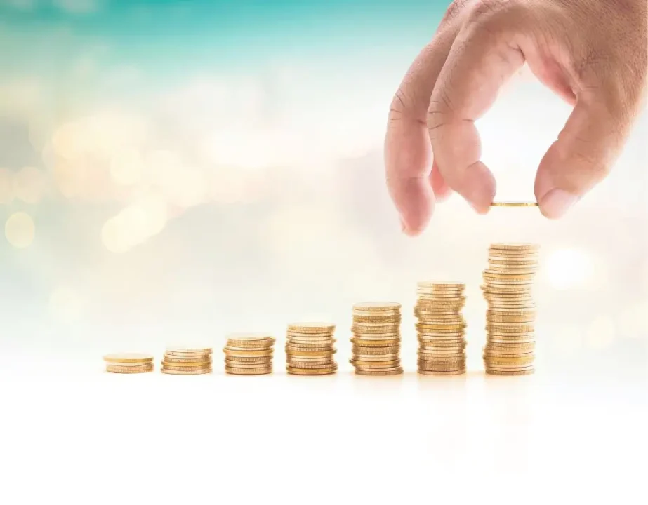 obrazek do artykułu - dofinansowanie wynagrodzeń PFRON. Obraz ukazuje stopniowo rosnące stosy złotych monet, symbolizujące akumulację kapitału lub wzrost finansowy. Nad najwyższym stosem widoczna jest ręka, która delikatnie dokłada monetę, co dodatkowo sugeruje proces inwestowania lub oszczędzania.