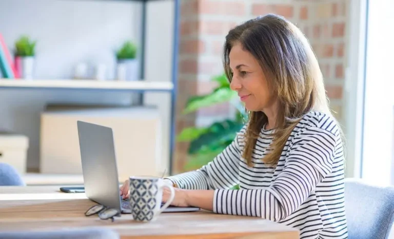 Na zdjęciu widzimy kobietę siedzącą przy stole i korzystającą z laptopa. Jest uśmiechnięta i wygląda na skoncentrowaną. Ubrana jest w biało-czarną bluzkę w paski. Na stole, obok laptopa, znajduje się filiżanka w biało-granatowy wzór, a obok niej okulary. W tle widać regał z książkami, ozdobami i roślinami doniczkowymi, co nadaje przestrzeni przytulny i domowy charakter. Zdjęcie może symbolizować pracę zdalną, naukę online lub uczestnictwo w sesjach psychoterapii online w spokojnym otoczeniu.