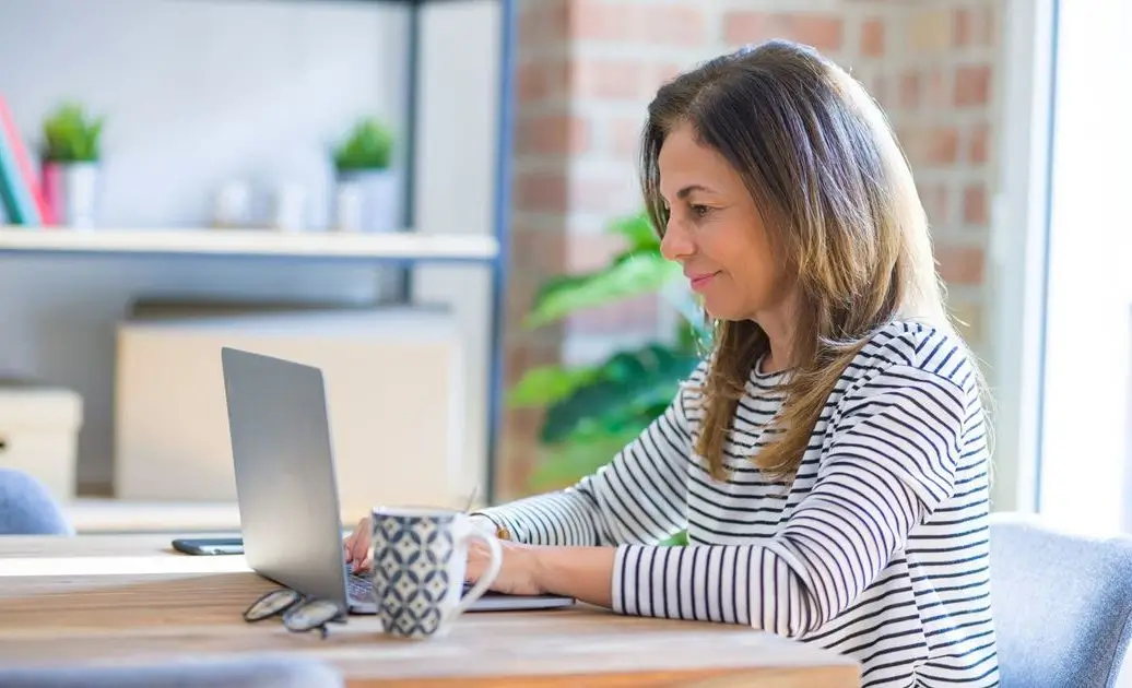 Na zdjęciu widzimy kobietę siedzącą przy stole i korzystającą z laptopa. Jest uśmiechnięta i wygląda na skoncentrowaną. Ubrana jest w biało-czarną bluzkę w paski. Na stole, obok laptopa, znajduje się filiżanka w biało-granatowy wzór, a obok niej okulary. W tle widać regał z książkami, ozdobami i roślinami doniczkowymi, co nadaje przestrzeni przytulny i domowy charakter. Zdjęcie może symbolizować pracę zdalną, naukę online lub uczestnictwo w sesjach psychoterapii online w spokojnym otoczeniu.