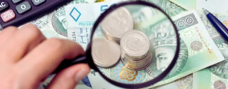 obrazek do artykułu - świadczenia dla niepełnosprawnych. Obraz przedstawia stos monet i polskie banknoty widziane przez lupę. Na pierwszym planie ręka trzyma lupę, co symbolizuje analizę finansową lub poszukiwanie oszczędności. W tle widoczne są elementy biurowe, takie jak długopis i kalkulator, co podkreśla kontekst związany z finansami.