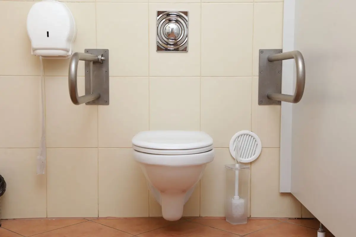 Zdjęcie przedstawia toaletę dostosowaną dla osób z niepełnosprawnościami. Centralnym elementem jest wisząca muszla klozetowa o białym kolorze, zamontowana na wysokości ułatwiającej korzystanie. Po obu stronach toalety znajdują się solidne, metalowe poręcze zamocowane do ściany, które mają pomóc w podtrzymaniu i przesiadaniu się. Ściany są wyłożone kremowymi, gładkimi płytkami ceramicznymi, co nadaje wnętrzu prosty i funkcjonalny wygląd. Na ścianie powyżej toalety znajduje się metalowy przycisk spłukujący o kwadratowym kształcie z zaokrąglonymi detalami. Po lewej stronie wisi biały podajnik na papier toaletowy o prostym, zaokrąglonym designie, a poniżej zwisa końcówka papieru. Na podłodze, po prawej stronie toalety, stoi przezroczysty pojemnik z plastikową szczotką do czyszczenia toalety. Podłoga jest wyłożona płytkami w ciepłym, ceglastym odcieniu, ułożonymi w skośny wzór. Pomieszczenie jest czyste, jasne i dostosowane do potrzeb osób wymagających dodatkowego wsparcia w korzystaniu z toalety.