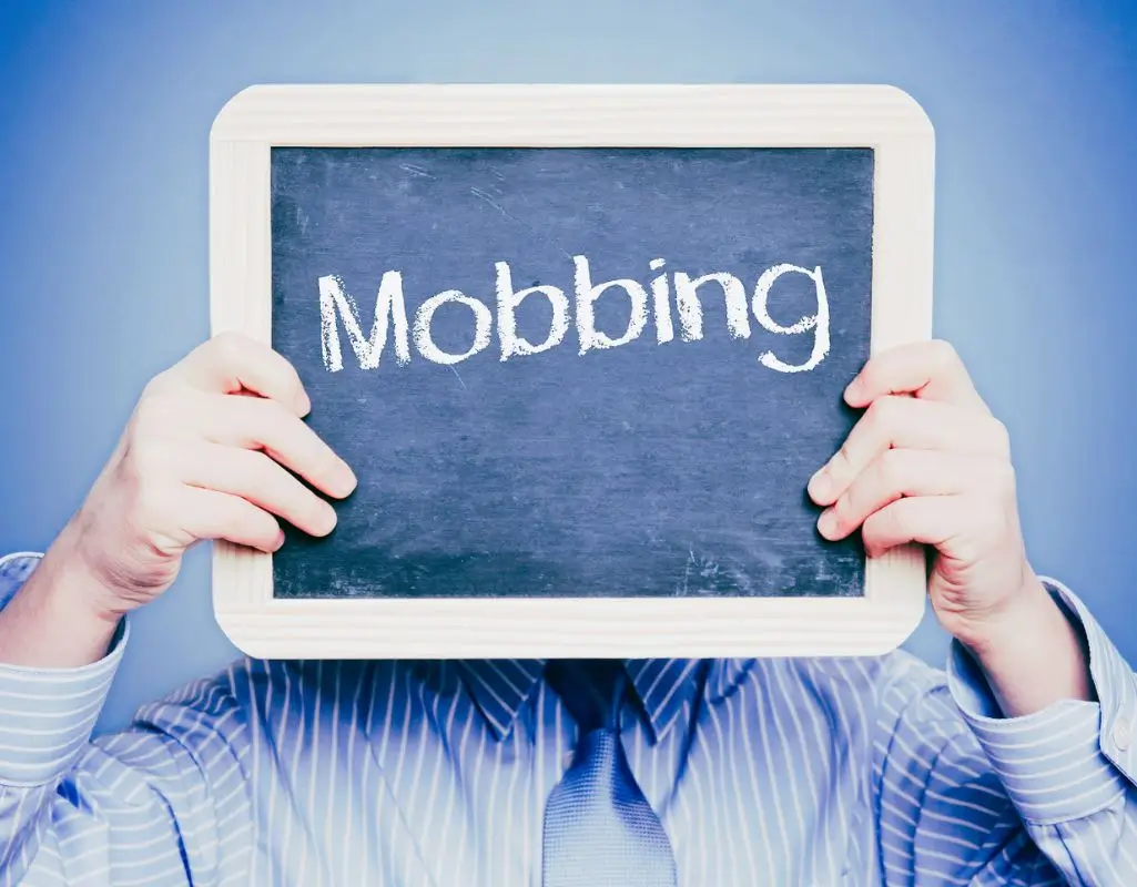 Na obrazku znajduje się osoba trzymająca tabliczkę w obu rękach na wysokości twarzy, na której widnieje napis „Mobbing” zapisany białą kredą na ciemnym tle tablicy. Postać jest ubrana w koszulę w delikatne niebieskie paski, co kontrastuje z intensywnie niebieskim tłem. Wyraz „Mobbing” centralnie umieszczony na tablicy przyciąga uwagę i wywołuje uczucia związane z dyskomfortem, napięciem i refleksją nad poważnym problemem społecznym, jakim jest mobbing w środowisku pracy. Obrazek sugeruje ukrywanie tożsamości lub próbę zwrócenia uwagi na problem, który często pozostaje niewidoczny. Sugeruje pytanie - jak sobie radzić z mobbingiem w pracy.