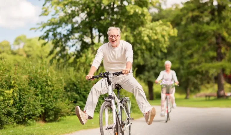 Zdjęcie przedstawia dwie starsze osoby, mężczyznę i kobietę, cieszące się przejażdżką na rowerach w słoneczny dzień. Na pierwszym planie znajduje się mężczyzna, który jedzie na rowerze z uniesionymi w górę nogami, wyraźnie bawiąc się i śmiejąc. Ubrany jest w jasny sweter i spodnie w kolorze beżowym, a na nogach ma wygodne, sportowe buty. Wyraz jego twarzy emanuje radością i beztroską. W tle, za mężczyzną, widać kobietę jadącą na rowerze z koszykiem pełnym kwiatów. Ubrana jest w jasne, pastelowe ubrania, które nadają scenie wesoły i lekki charakter. Oboje znajdują się na asfaltowej ścieżce otoczonej bujną zielenią – widać drzewa, krzewy i trawę, co sugeruje, że jest to park lub teren rekreacyjny. Atmosfera zdjęcia jest pełna energii, swobody i radości, ukazując aktywne spędzanie czasu na świeżym powietrzu. Idealnie oddaje chwilę pełną relaksu i zabawy.