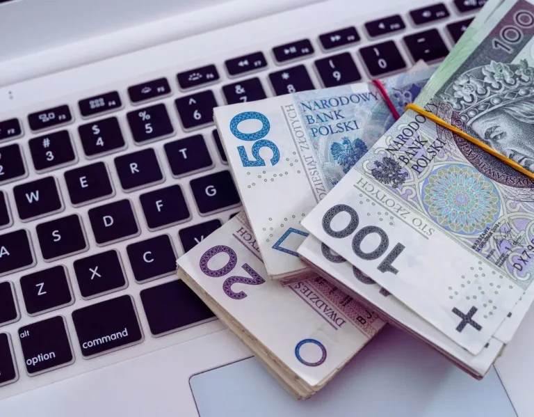 Obrazek do artykułu dofinansowanie wynagrodzeń PFRON w 2025. Na zdjęciu widać polskie banknoty w nominałach 20, 50 i 100 złotych, ułożone w stosy na klawiaturze laptopa. Ostre, wyraziste kolory pieniędzy kontrastują z czarnymi klawiszami, co może wzbudzać skojarzenia z przepływem gotówki i możliwościami finansowymi.