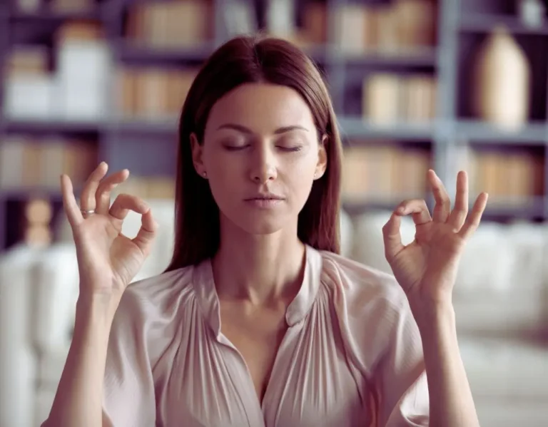 Na obrazie znajduje się kobieta z zamkniętymi oczami, siedząca w spokojnym otoczeniu. Jej dłonie są uniesione na wysokość ramion, z palcami ułożonymi w klasycznym geście medytacyjnym, co sugeruje, że praktykuje techniki relaksacyjne. Kobieta wygląda na zrelaksowaną, skoncentrowaną i wyciszoną, co odzwierciedla spokój ducha i harmonię. Tło jest rozmyte, co podkreśla intymność i wewnętrzne skupienie. Ten obraz idealnie ilustruje, jak radzić sobie ze stresem poprzez medytację i praktyki mindfulness.