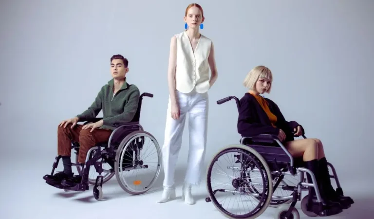 Na zdjęciu widzimy trójkę osób, co może symbolizować różne stopnie niepełnosprawności i indywidualne podejście do życia z ograniczeniami. Dwie osoby na wózkach inwalidzkich reprezentują różne potrzeby wynikające z ograniczeń ruchowych – mogą one odnosić się do stopnia umiarkowanego lub znacznego, zależnie od zakresu samodzielności. Mężczyzna ubrany w sweter i kobieta w czarnej bluzie pokazują, że mimo pewnych trudności, osoby z niepełnosprawnością mogą wyrażać swoją osobowość i styl. Kobieta stojąca pośrodku może symbolizować osobę wspierającą, ale również zwraca uwagę na to, że stopnie niepełnosprawności mogą być niewidoczne na pierwszy rzut oka, np. w przypadku chorób przewlekłych czy schorzeń ukrytych.