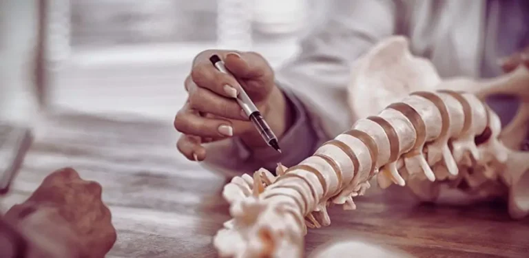 Na obrazku widzimy zbliżenie na model ludzkiego kręgosłupa, który jest badany przez specjalistę. Osoba w białym fartuchu, prawdopodobnie lekarz lub fizjoterapeuta, trzyma długopis i wskazuje nim na konkretne miejsce na modelu. W tle widoczna jest rozmyta sylwetka pacjenta, co sugeruje konsultację medyczną. Zdjęcie utrzymane jest w ciepłej tonacji, co nadaje mu spokojny, profesjonalny charakter. Ukazuje ono precyzję i troskę w analizie problemów zdrowotnych związanych z kręgosłupem, podkreślając znaczenie specjalistycznej diagnozy. Obraz dobrze ilustruje tematykę związaną z pytaniem - czy na kręgosłup można dostać orzeczenie o niepełnosprawności - i niesie ze sobą skojarzenia z medycznym wsparciem oraz poszukiwaniem ulgi w bólu.