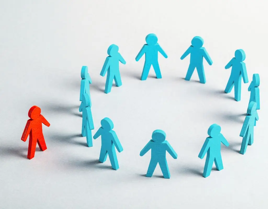 Czym jest dyskryminacja w pracy, jak ją identyfikować oraz jej przeciwdziałać? 1 Na obrazku przedstawiono grupę niebieskich figur symbolizujących ludzi, ustawionych w kręgu. Na zewnątrz, wyraźnie odizolowana, znajduje się czerwona figurka, która wydaje się wykluczona z grupy. Układ postaci sugeruje dystans, odrzucenie i brak akceptacji. Kompozycja wizualnie oddaje poczucie izolacji, samotności i nierównego traktowania, co może nawiązywać do problemu społecznego, jakim jest wykluczenie. W kontekście pytania - czym jest dyskryminacja w pracy - obraz może symbolizować sytuację, w której jednostka jest celowo pomijana, niesprawiedliwie traktowana lub marginalizowana ze względu na cechy, które ją wyróżniają.