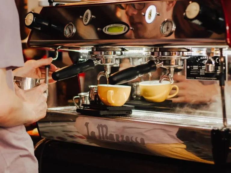 Zdjęcie przedstawia proces parzenia kawy w kawiarni, ukazując profesjonalny ekspres do kawy marki La Marzocco. Barista, którego twarz odbija się w lustrzanej powierzchni ekspresu, przygotowuje kawę, trzymając metalowy dzbanek do spieniania mleka, prawdopodobnie do przyrządzenia cappuccino lub latte. Szczegóły obrazu: • Ekspres do kawy: Duży, profesjonalny ekspres kolbowy z dźwigniami, wskaźnikami ciśnienia i połyskującą metalową obudową, odbijającą otoczenie. • Filiżanki: Dwie żółte filiżanki stoją na czarnych podstawkach, a z kolby ekspresu leje się aromatyczna kawa. • Barista: Widoczny w odbiciu maszyny, skupiony na przygotowaniu kawy. Jego ręce trzymają dzbanek do spieniania mleka, co sugeruje, że przygotowuje napój mleczny. • Ciepła atmosfera: Oświetlenie jest przytulne, dominują ciepłe odcienie brązu i złota, co oddaje klimat małej, rzemieślniczej kawiarni. Zdjęcie doskonale oddaje klimat pracy baristy i pasję do kawy.