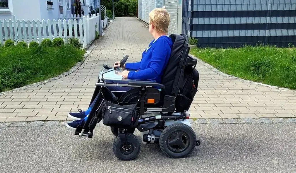 Osoba na wózku inwalidzkim w ruchu pieszym i transporcie. Uprawnienia i zasady 1 Na zdjęciu widoczna jest osoba na wózku inwalidzkim, ubrana w niebieski strój, która porusza się po spokojnej, wyłożonej płytkami uliczce. W tle znajdują się domy otoczone białymi płotami oraz zielone trawniki. Osoba na wózku inwalidzkim wygląda na spokojną i skupioną, patrząc w stronę drogi, co może sugerować poczucie niezależności i swobody. Zdjęcie oddaje atmosferę cichej, przyjaznej okolicy, która wydaje się przystosowana do potrzeb osób z ograniczeniami ruchowymi.