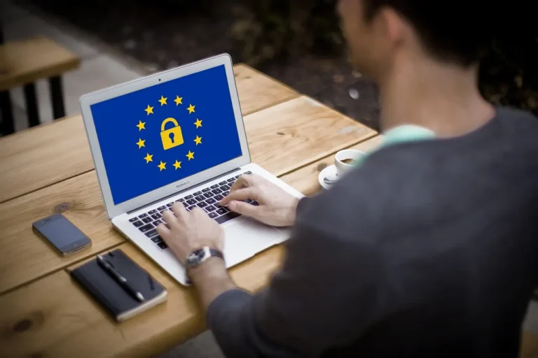 Wyjaśniamy, jak spełnić wymogi RODO w mailach transakcyjnych 6 Zdjęcie przedstawia mężczyznę pracującego na laptopie, siedzącego przy drewnianym stole na świeżym powietrzu. Na ekranie komputera widoczna jest grafika symbolizująca ochronę danych w Unii Europejskiej, przedstawiająca żółtą kłódkę otoczoną dwunastoma gwiazdami na niebieskim tle, nawiązującą do flagi UE. Szczegóły obrazu: • Mężczyzna: • Siedzi przy drewnianym stole, ujęcie jest od tyłu, więc jego twarz nie jest widoczna. • Ma na ręku zegarek, co sugeruje dbałość o czas i organizację pracy. • Laptop: • Srebrny, prawdopodobnie MacBook Air, używany przez mężczyznę do pracy lub przeglądania informacji. • Na ekranie grafika nawiązująca do RODO (GDPR) – europejskiego prawa o ochronie danych. • Otoczenie: • Drewniany stół na świeżym powietrzu, co może sugerować pracę zdalną lub freelancerstwo. • Na stole znajdują się: • Smartfon, co sugeruje wielozadaniowość użytkownika. • Czarna skórzana notatnik i długopis, dodające profesjonalnego charakteru. • Filiżanka kawy na białym spodku, co podkreśla atmosferę relaksu i pracy jednocześnie. Atmosfera i przekaz: Zdjęcie symbolizuje cyfrową prywatność, ochronę danych osobowych oraz pracę w zgodzie z przepisami UE. Może być używane w kontekście tematów takich jak bezpieczeństwo w sieci, RODO, cyberbezpieczeństwo czy praca zdalna.