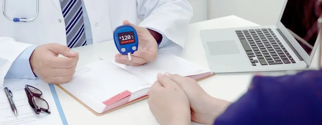 Na zdjęciu widać lekarza w białym fartuchu, który trzyma glukometr wskazujący poziom cukru (120 mg/dL) i omawia wyniki z pacjentką siedzącą naprzeciw. Na biurku obok leży laptop oraz notatnik. Atmosfera sugeruje troskę i chęć wsparcia, ale też odrobinę niepewności u pacjentki. Towarzyszy temu pytanie „Czy na cukrzycę można dostać orzeczenie”, które podkreśla zarówno potrzebę zrozumienia sytuacji zdrowotnej, jak i nadzieję na uzyskanie konkretnej pomocy.