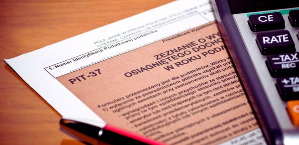 Na zdjęciu znajduje się formularz PIT-37, częściowo wsunięty w białą teczkę, leżący na drewnianym biurku. Na pierwszym planie widoczny jest fragment kalkulatora z przyciskami oznaczonymi m.in. "TAX", "RATE" i "CE", co sugeruje obliczenia podatkowe. W tle długopis, gotowy do wypełnienia dokumentów. Zdjęcie oddaje atmosferę corocznego rozliczenia podatkowego – mieszankę skupienia, odpowiedzialności i niepewności co do finalnej kwoty zwrotu. Hasło "ulga rehabilitacyjna 2280 zł. Ile zwrotu łącznie zyskasz?" idealnie pasuje do kontekstu, skłaniając do refleksji nad możliwymi oszczędnościami i zachęcając do sprawdzenia swoich uprawnień podatkowych.