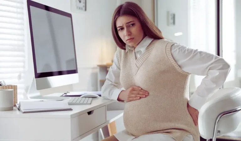 Kobieta w zaawansowanej ciąży siedzi przy biurku w jasnym, biurowym wnętrzu. Ubrana w białą koszulę i beżowy sweter, z troską i dyskomfortem trzyma dłoń na brzuchu oraz podpierając plecy – sygnalizując ból i zmęczenie związane z ciążą. Wyraz jej twarzy sugeruje niepewność i potrzebę dodatkowego wsparcia w codziennych obowiązkach. Sytuacja na zdjęciu przywodzi na myśl potrzebę odpoczynku oraz rozważenia rozwiązań takich jak uzupełniający urlop macierzyński.