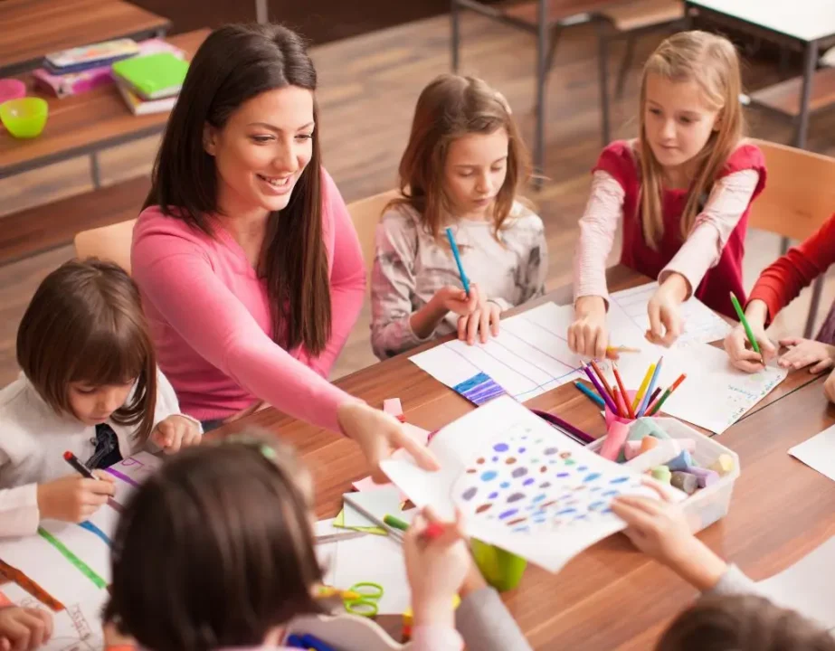 Zdjęcie przedstawia grupę dzieci oraz młodą nauczycielkę w trakcie zajęć plastycznych w klasie. W centrum kadru siedzi uśmiechnięta kobieta o długich, brązowych włosach, ubrana w różowy sweter. Wspólnie z dziećmi pracuje przy dużym, drewnianym stole. Wokół niej znajduje się sześcioro dzieci w wieku wczesnoszkolnym, zarówno dziewczynki, jak i chłopcy, zaangażowane w rysowanie i kolorowanie. Dzieci używają kolorowych pisaków i kredek. Na stole rozłożone są arkusze papieru z różnorodnymi rysunkami, pudełko z kredą, temperówką i innymi przyborami szkolnymi. W prawym dolnym rogu widać ręce dziecka trzymającego kartkę z kolorowymi plamkami farby lub flamastra. W tle widoczne są drewniane ławki i krzesła, sugerujące, że zdjęcie wykonano w sali lekcyjnej. Atmosfera sceny jest ciepła, radosna i kreatywna — obraz oddaje moment aktywnej zabawy i nauki poprzez sztukę.