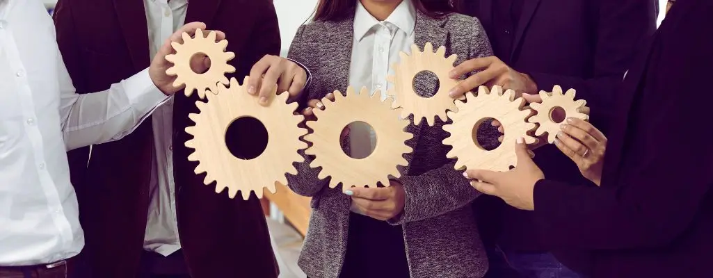 Na zdjęciu widać grupę osób ubranych w formalne stroje biznesowe, które wspólnie trzymają w rękach drewniane zębatki. Każda osoba odpowiada za inny element mechanizmu, a wszystkie koła są ze sobą połączone, co symbolizuje współpracę, wzajemne wsparcie i wspólne dążenie do celu. Obrazek oddaje poczucie zaangażowania, zgrania zespołu i odpowiedzialności. Pokazuje, że kiedy każda jednostka wnosi swoje umiejętności i działa zgodnie z resztą grupy, całość funkcjonuje jak dobrze naoliwiona maszyna. To wizualna metafora oddająca cechy dobrego pracownika – takich jak współpraca, zaangażowanie, odpowiedzialność, komunikatywność i gotowość do działania w zespole.