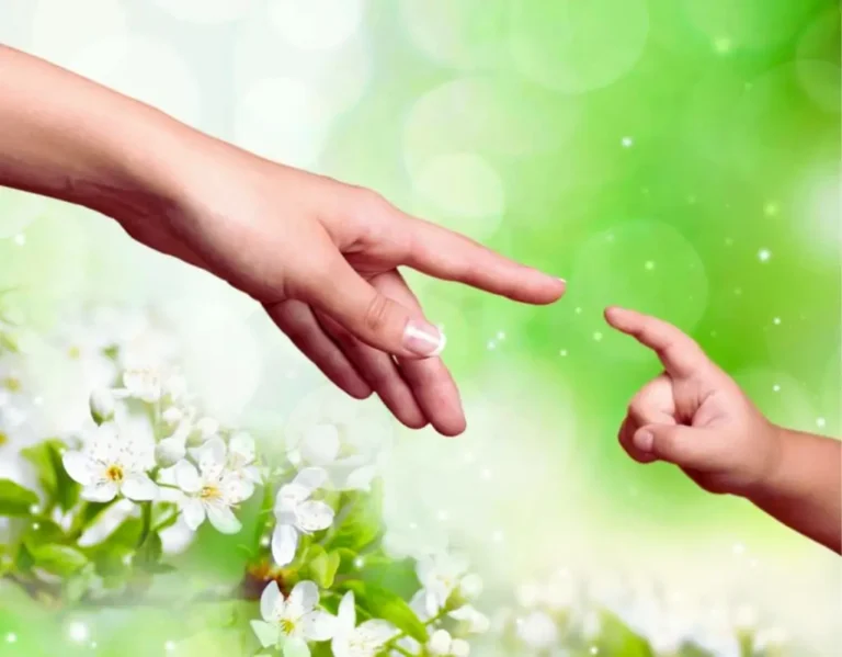 Na obrazku widzimy delikatny gest zbliżających się do siebie dłoni – dorosłej osoby i małego dziecka. Palce niemal się dotykają, co przywodzi na myśl więź, bliskość i wzajemne zaufanie. W tle znajdują się rozmyte, zielone liście i białe kwiaty, tworzące wiosenną, spokojną atmosferę. Całość symbolizuje miłość, opiekę i dobre relacje z dzieckiem – to moment porozumienia bez słów, pełen czułości i bezpieczeństwa.