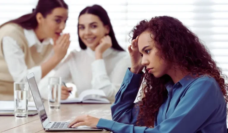 Na zdjęciu widać trzy kobiety w biurowym otoczeniu. Na pierwszym planie siedzi młoda kobieta o kręconych włosach, która wygląda na przygnębioną i zaniepokojoną. Pracuje przy laptopie, opierając głowę na ręce. W tle, dwie inne kobiety szeptają do siebie i śmieją się, spoglądając w jej stronę. Ich zachowanie sugeruje plotkowanie lub komentowanie jej osoby. Obraz oddaje emocje samotności, wykluczenia i stresu, które mogą być skutkiem sytuacji, jaką jest dyskryminacja pośrednia w pracy.