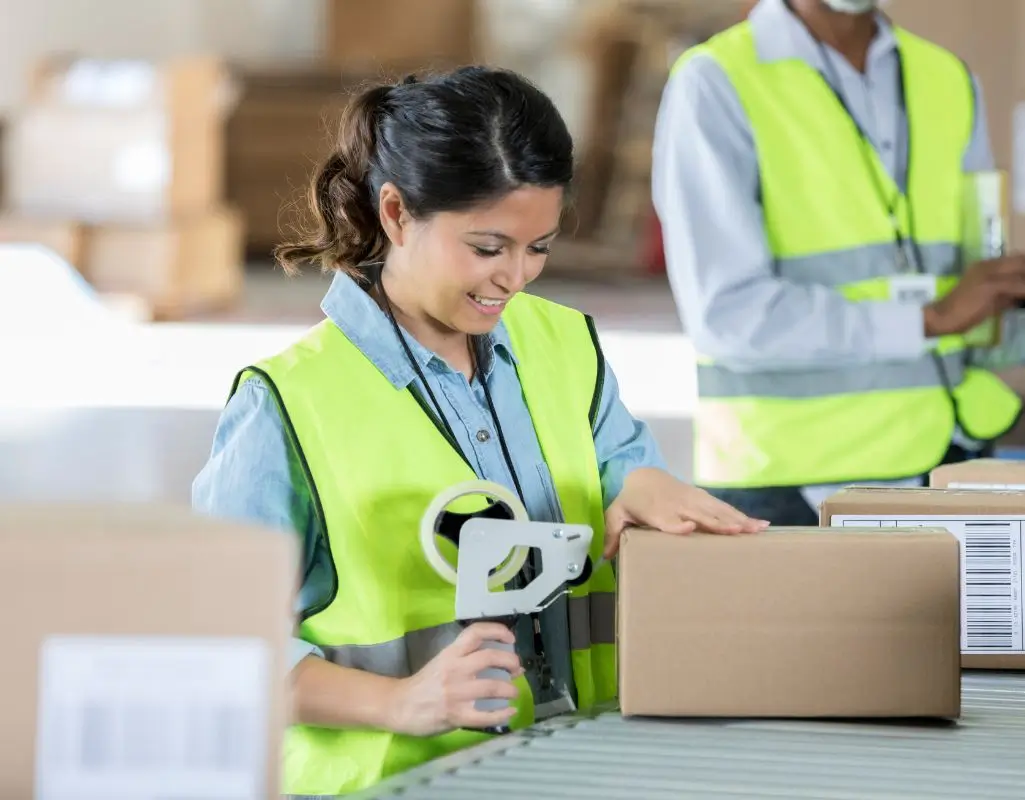 Uśmiechnięta kobieta w odblaskowej, żółtej kamizelce zabezpieczającej pakuje kartonowe pudełko przy taśmie produkcyjnej. W jednej ręce trzyma dyspenser do taśmy klejącej, a drugą ręką przytrzymuje paczkę. W tle znajduje się inny pracownik w podobnej kamizelce, a wokół widoczne są inne kartony i elementy hali magazynowej lub sortowni.