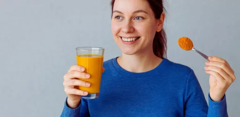 Czy kurkuma pomaga na depresję i co warto wiedzieć, zanim ją wypróbujesz? 2 Na obrazku znajduje się uśmiechnięta kobieta w niebieskiej bluzie, trzymająca w jednej ręce szklankę z napojem o intensywnie żółtopomarańczowym kolorze, a w drugiej łyżeczkę z mieloną kurkumą. Jej wyraz twarzy sugeruje pozytywne emocje – wygląda na zadowoloną i pełną nadziei. Scena oddaje atmosferę troski o zdrowie, naturalnego podejścia do samopoczucia i świadomego wyboru. Obrazek może kojarzyć się z pytaniem: czy kurkuma pomaga w depresji, i sugeruje, że sięganie po naturalne środki może być elementem dbania o dobre samopoczucie.