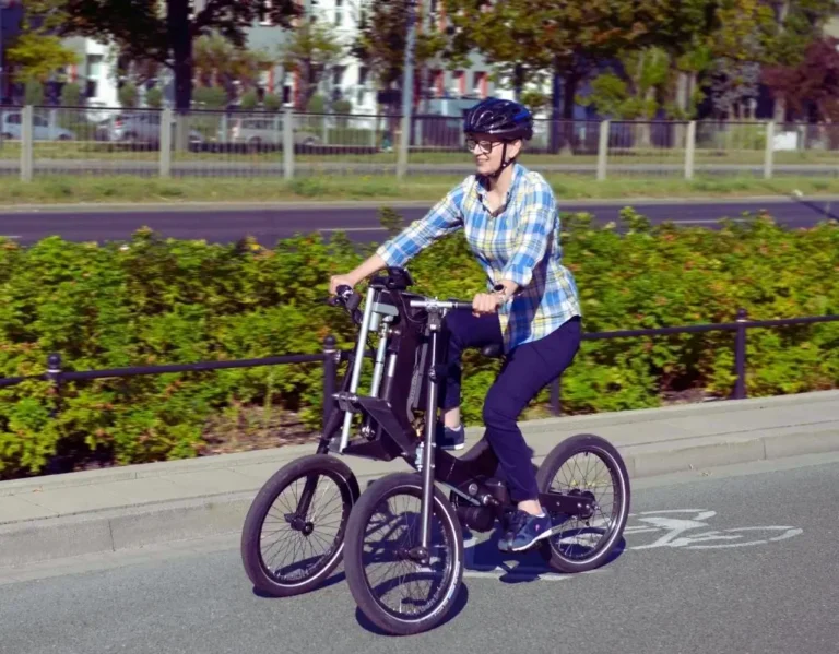 Na zdjęciu widać uśmiechniętą kobietę jadącą nowoczesnym, elektrycznym rowerem trójkołowym po ścieżce rowerowej w słoneczny dzień. Ma na sobie kask i sportowe ubranie, co podkreśla aktywny tryb życia. Rower, który prowadzi, posiada nietypową, stabilną konstrukcję, idealną dla osób z ograniczoną mobilnością. W tle widać zieleń miejską i zabudowania, co sugeruje, że jazda odbywa się w przyjaznym, miejskim otoczeniu. Obrazek oddaje poczucie wolności, niezależności i radości z ruchu na świeżym powietrzu – doświadczenie, które może umożliwić dofinansowanie do roweru elektrycznego z PFRON.