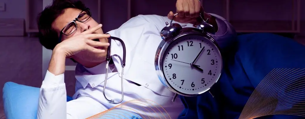 Na zdjęciu widać zmęczonego mężczyznę w kitlu lekarskim, który leży w łóżku i ziewa, trzymając w ręku duży budzik pokazujący godzinę 4:55. Ma na szyi stetoskop i okulary, co sugeruje, że jest lekarzem lub pracownikiem medycznym. Jego twarz wyraża wyczerpanie i senność, typowe dla osób pracujących w nocy. Obraz oddaje trud i dyskomfort związany z pracą na nocnej zmianie, ilustrując temat – jak radzić sobie z nocną zmianą – poprzez zmęczenie, brak snu i konieczność funkcjonowania w nietypowych godzinach.