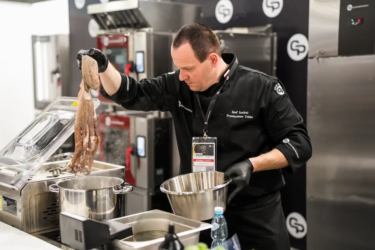Szef kuchni w profesjonalnej kuchni gastronomicznej przygotowuje ośmiornicę, trzymając ją nad garnkiem z wrzątkiem. W tle widoczne nowoczesne piece konwekcyjno-parowe i sprzęt kuchenny. Obraz ukazuje zaawansowane wyposażenie gastronomiczne w akcji – symbol precyzyjnego gotowania i nowoczesnej technologii w kuchni.