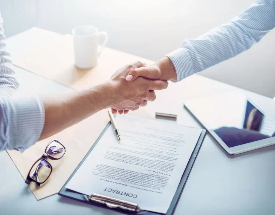 Jak rozwiązać umowę o pracę za porozumieniem stron. Poradnik krok po kroku ze wzorem 2 Na zdjęciu widać dwie osoby uściślające sobie dłonie nad dokumentem leżącym na biurku – to symbol zakończenia formalnych ustaleń i wzajemnego porozumienia. Obok znajduje się długopis, tablet oraz okulary, co podkreśla biurowy, profesjonalny charakter sytuacji. Jasne światło i uporządkowana przestrzeń budują atmosferę spokoju i wzajemnego szacunku. Scena doskonale ilustruje moment, w którym następuje rozwiązanie umowy o pracę za porozumieniem stron – spokojne, świadome i zgodne rozstanie dwóch stron współpracy.