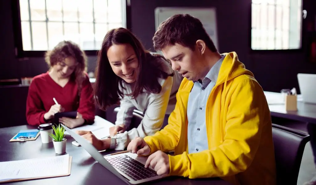 Na zdjęciu widać uczestników zajęć. Młody mężczyzna w żółtej bluzie pracuje przy laptopie, a uśmiechnięta kobieta wspiera go i dodaje otuchy. W tle inna uczestniczka notuje coś w zeszycie. Cała scena pokazuje atmosferę życzliwości, zaangażowania i wspólnej nauki, jaką daje Warsztat Terapii Zajęciowej.