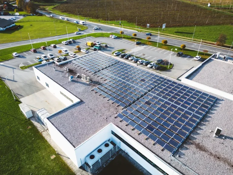 Jak zmniejszyć wydatki na flotę firmową i zaoszczędzić tysiące złotych? 8 Widok z lotu ptaka na nowoczesny budynek firmowy z dużą instalacją paneli fotowoltaicznych na dachu i parkingiem pełnym samochodów. Obraz symbolizuje inwestycję w odnawialne źródła energii jako sposób na redukcję kosztów eksploatacji, w tym zasilanie floty elektrycznej tańszym, własnym prądem