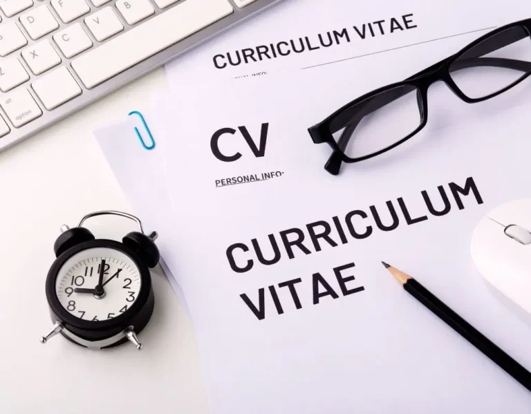 Na zdjęciu znajduje się jasne biurko, na którym leżą dokumenty - wzór jak napisać CV. Obok dokumentów są czarne okulary, ołówek, białą myszkę oraz fragment klawiatury.
