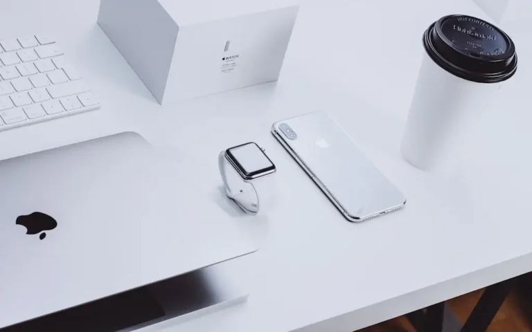 Nowoczesne biurko z urządzeniami Apple: zamknięty MacBook, Apple Watch na białym pasku, iPhone w srebrnej obudowie, klawiatura oraz kubek z kawą na wynos. Obraz symbolizuje elegancję, funkcjonalność i spójność ekosystemu Apple – technologii zaprojektowanej z myślą o dostępności i codziennym komforcie użytkownika.