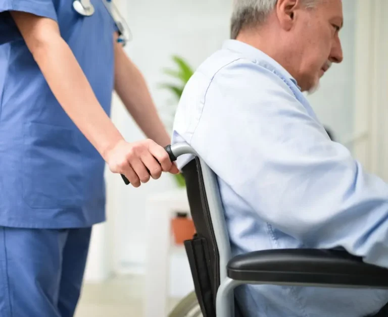 Starszy mężczyzna na wózku inwalidzkim siedzi bokiem, prowadzony przez osobę w niebieskim stroju medycznym – symbol wsparcia i opieki w kontekście podejmowania trudnych decyzji prawnych dotyczących bliskich z niepełnosprawnością.
