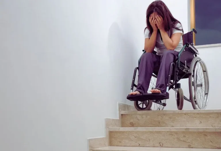 Na zdjęciu widać młodą kobietę na wózku inwalidzkim, siedzącą na szczycie schodów w mieszkaniu. Kobieta zasłania twarz dłońmi, wyrażając bezradność, smutek i frustrację. Jej pozycja i otoczenie podkreślają brak dostępu i wykluczenie z przestrzeni, która nie została przystosowana do potrzeb osób z niepełnosprawnością. Obraz silnie pokazuje, jak ważna jest likwidacja barier architektonicznych z dofinansowaniem PFRON – by uniknąć takich sytuacji i dać każdemu szansę na niezależność i godność.