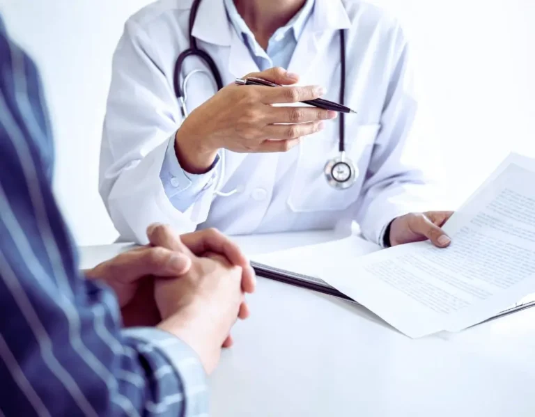 Na zdjęciu widać lekarza w białym fartuchu, ze stetoskopem na szyi, który rozmawia z pacjentem. Trzyma dokumenty i długopis, wyjaśniając coś z zaangażowaniem. Pacjent siedzi naprzeciwko, ze splecionymi dłońmi, wsłuchany w to, co mówi specjalista. To moment pełen skupienia i emocji, często towarzyszący rozmowom o stanie zdrowia i świadczeniach, takich jak najczęściej przyznawane renty.