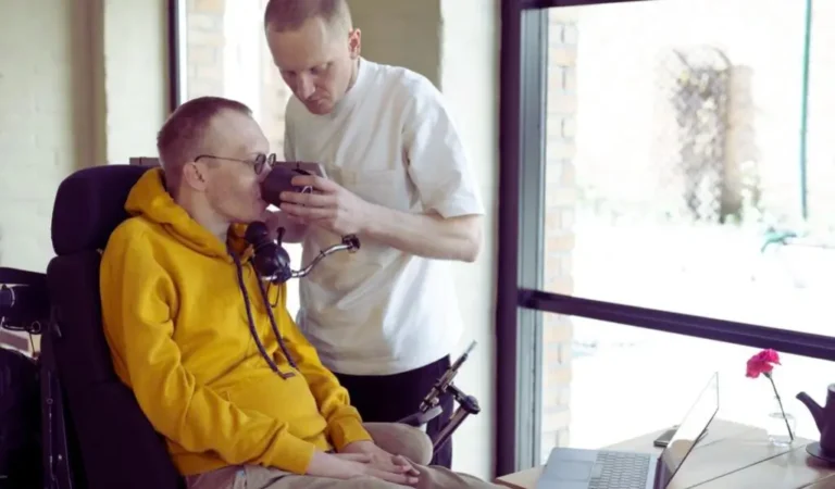 Na zdjęciu widać dwóch mężczyzn w jasnym, przytulnym wnętrzu. Jeden z nich, osoba z tetraplegią, siedzi na wózku inwalidzkim i ma na sobie żółtą bluzę z kapturem. Korzysta z urządzenia wspomagającego komunikację zamontowanego przy twarzy. Drugi mężczyzna, ubrany w białą koszulkę, z troską i skupieniem podaje mu kubek z napojem, pomagając napić się. W ich spojrzeniach i gestach widać zaufanie, empatię i codzienną bliskość. Obraz ukazuje nie tylko wyzwania związane z porażeniem 4 kończyn (tetraplegia), ale przede wszystkim siłę relacji międzyludzkich i znaczenie wsparcia w codziennym życiu.