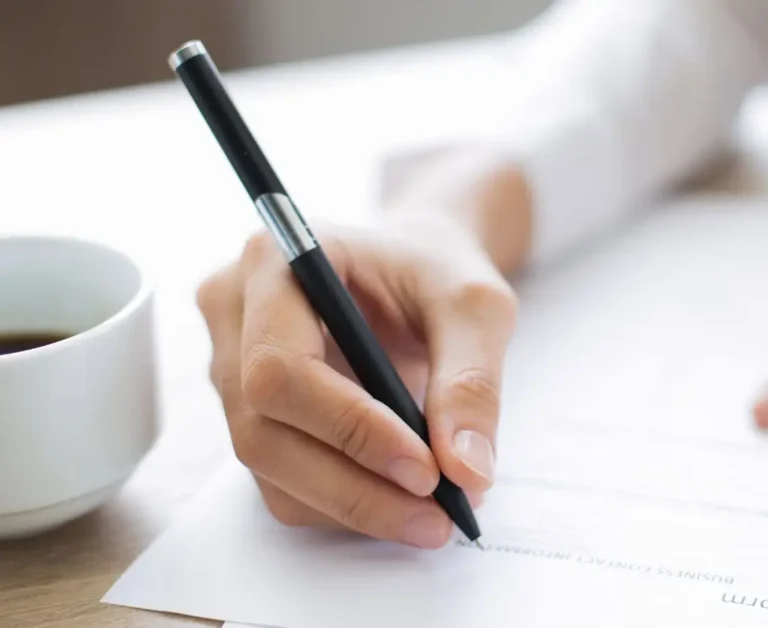 Zbliżenie na dłoń osoby podpisującej dokument czarnym długopisem – na kartce widoczny fragment formularza. Po lewej stronie stoi filiżanka z kawą. Obraz ilustruje proces składania lub zatwierdzania wniosków, symbolizując formalności związane z zarządzaniem dokumentacją urlopową w organizacjach.