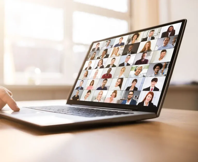 Laptop na biurku z wyświetlonym ekranem spotkania online, pokazującym kilkadziesiąt twarzy uczestników w trybie wideokonferencji. Obraz symbolizuje prowadzenie webinarów i szkoleń online – nawet dla kameralnej, ale zaangażowanej grupy odbiorców.