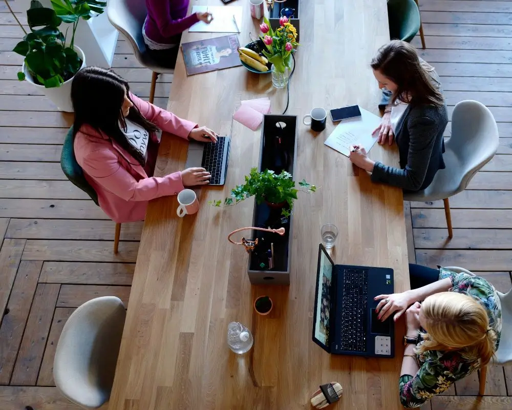 Widok z góry na elegancki drewniany stół w nowoczesnym, wspólnym biurze typu coworking. Przy stole pracują cztery kobiety — dwie na laptopach, jedna pisze notatki, a jedna przegląda magazyn. Na stole stoją kubki, karafka z wodą, laptop, świeże kwiaty i miska z owocami. Estetyczne, zielone wnętrze symbolizuje nowoczesne, elastyczne miejsca pracy — idealne do prowadzenia działalności pod adresem wirtualnym.