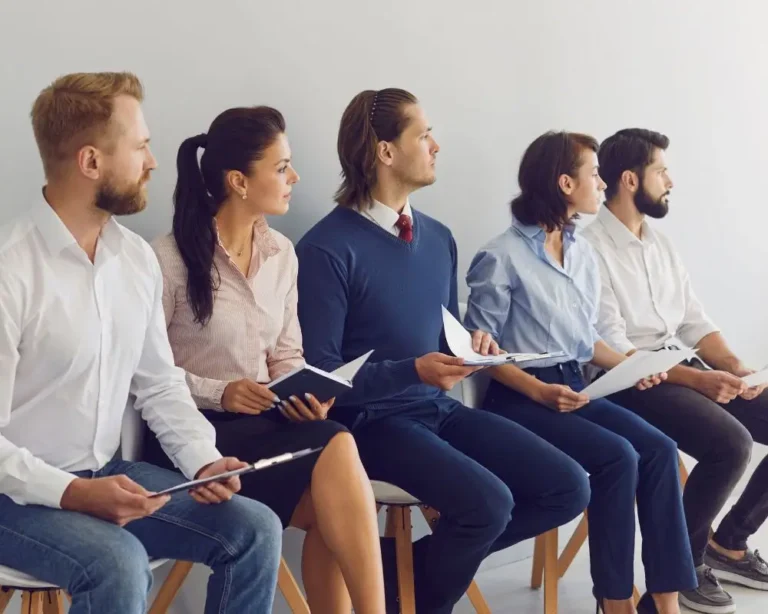 Pięć osób – trzech mężczyzn i dwie kobiety – siedzi w jednym rzędzie na krzesłach w poczekalni, każda z dokumentami w ręku (CV, teczki, notatniki). Wszyscy są elegancko ubrani i patrzą w tym samym kierunku, jakby wyczekiwali na rozmowę kwalifikacyjną. Zdjęcie symbolizuje proces rekrutacji, konkurencję na rynku pracy i potrzebę profesjonalnego wsparcia, jakie oferuje agencja rekrutacyjna.