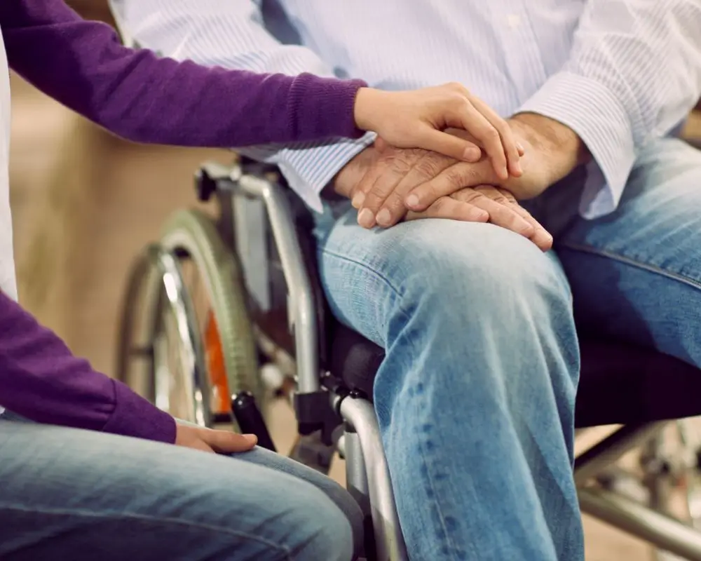 Jak uzyskać alimenty na dorosłe dziecko z niepełnosprawnością? 1 Zbliżenie na ręce dwóch osób – młodsza osoba w bluzie kładzie dłoń wspierająco na dłoni starszego mężczyzny siedzącego na wózku inwalidzkim, ubranego w dżinsy i koszulę. Symboliczna scena troski, bliskości i opieki w kontekście dorosłego dziecka z niepełnosprawnością, które mimo pełnoletniości wciąż wymaga wsparcia i alimentów.