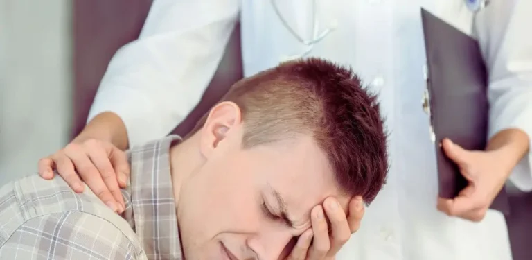 Na zdjęciu widać młodego mężczyznę, który z wyraźnym przygnębieniem trzyma głowę w dłoniach — jego twarz zdradza zmęczenie psychiczne i wewnętrzne cierpienie. Za nim stoi osoba w białym fartuchu lekarskim, trzymająca dokumentację medyczną i wspierająco kładąca dłoń na jego ramieniu. To poruszająca scena, pełna empatii i zrozumienia, która symbolicznie pokazuje, z czym mierzą się osoby w kryzysie psychicznym. Obraz dobrze oddaje emocje towarzyszące pytaniu, które zadaje sobie wielu pacjentów: czy na depresję można dostać orzeczenie o niepełnosprawności.