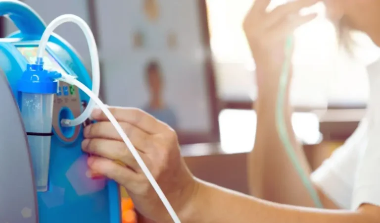 Na zdjęciu widać osobę korzystającą z koncentratora tlenu – urządzenia, które pomaga swobodniej oddychać w warunkach domowych. Delikatne światło i skupienie na dłoniach podłączających rurkę tworzą atmosferę codziennej troski o zdrowie i poczucia bezpieczeństwa. To cichy moment ulgi i nadziei, jaki niesie ze sobą dostęp do niezbędnego wsparcia. W takich sytuacjach dodatek PFRON do energii elektrycznej może odegrać kluczową rolę – dzięki niemu osoby z niepełnosprawnościami mogą bez obaw korzystać z urządzeń medycznych, które zużywają dużo prądu, a jednocześnie są niezbędne do życia.