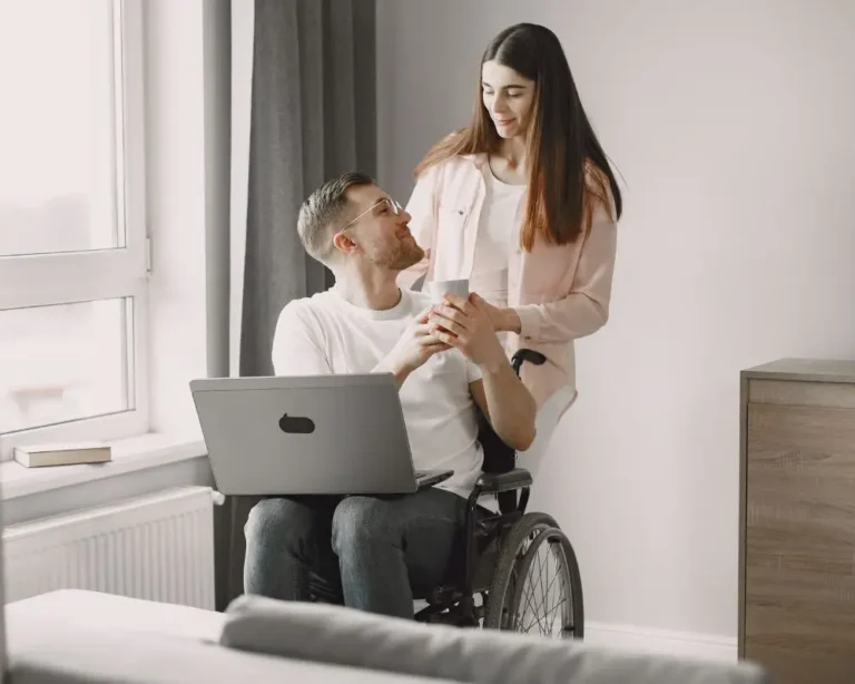 Jak budować intymność pomimo ograniczeń ruchowych? 12 Mężczyzna na wózku inwalidzkim siedzi przy oknie z laptopem na kolanach i patrzy z uśmiechem na stojącą obok młodą kobietę, która delikatnie trzyma jego dłonie i telefon. Para patrzy sobie w oczy z czułością. Otoczenie jest jasne i przytulne – białe ściany, zasłony, komoda, parapet z książką. Zdjęcie symbolizuje bliskość, zrozumienie i emocjonalne połączenie między partnerami, mimo fizycznych barier.