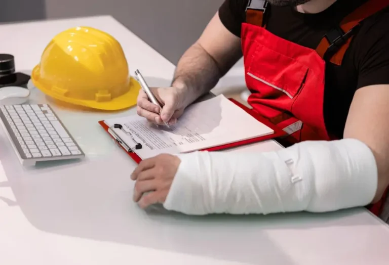 Mężczyzna w roboczym stroju, z ręką w gipsie, siedzi przy biurku i wypełnia dokumenty, prawdopodobnie związane z wypadkiem w pracy. Obok leży żółty kask ochronny, a na biurku znajduje się klawiatura i teczka z formularzami. Cała scena oddaje moment starania się o odszkodowanie z ZUS za wypadek w pracy – widać skupienie, zmęczenie i próbę uporania się z formalnościami mimo urazu. Obraz pokazuje nie tylko fizyczną kontuzję, ale też emocjonalne obciążenie i determinację, by odzyskać sprawiedliwość i wsparcie po trudnym doświadczeniu.